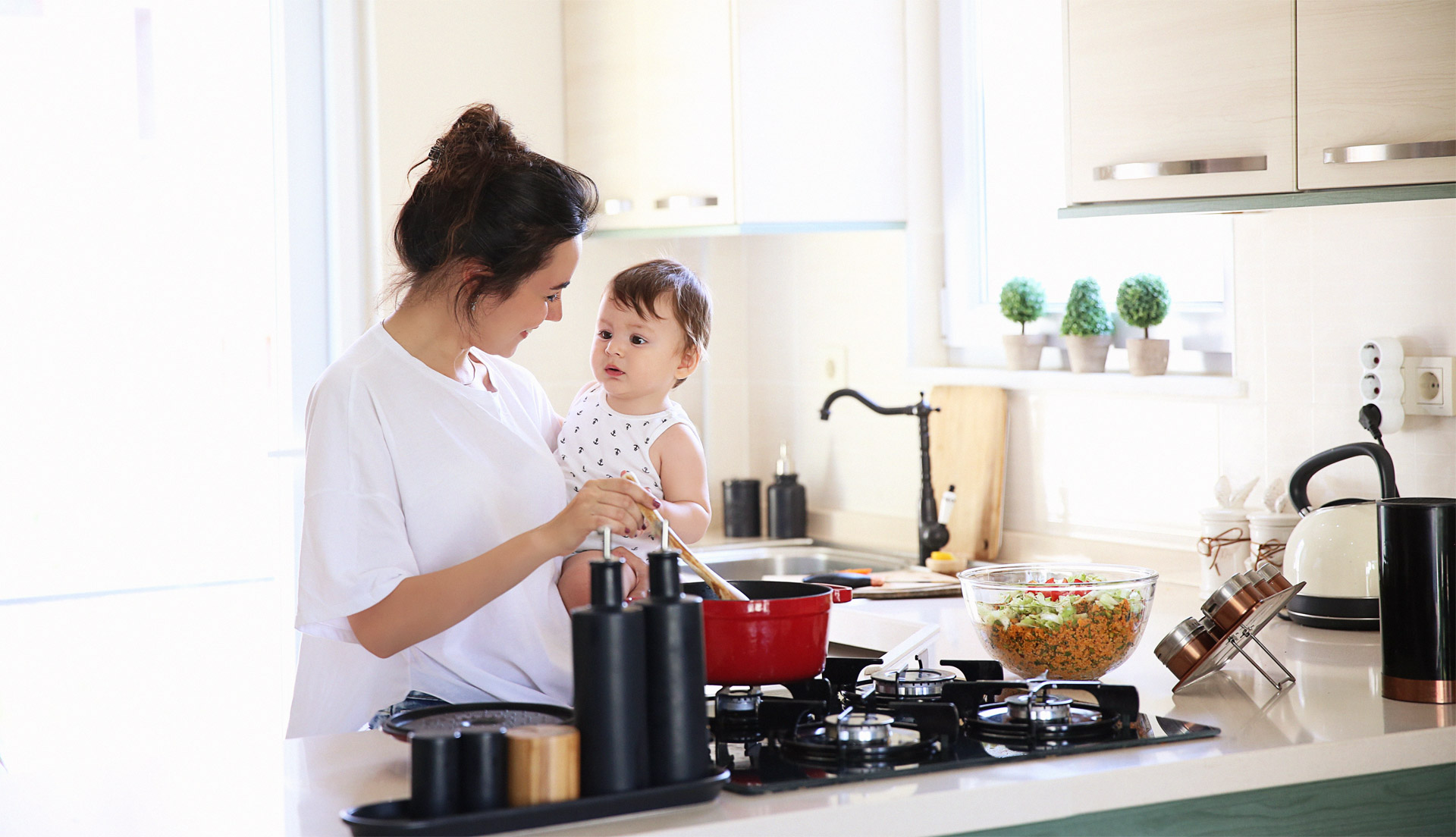 A mother holding a toddler and cooking food